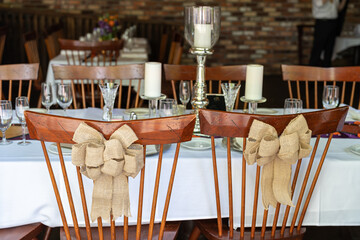 Two wooden seats with bows adorned on the back of them are tucked under a table at a wedding. The reception area is still being set up so all the seating is clean. 