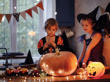 Little Sisters In Witch Costume Playing With Glowing Jack-o-lantern During Halloween Celebration In Dark Kitchen Room, Excited Kids And Pumpkins On Table