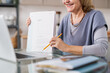 © pressmaster - Happy mature female showing document with financial information while sitting in front of laptop during online course of study