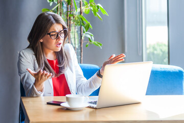 Unhappy woman employee in glasses raising hands in indignant gesture, asking why and looking angrily, looking at laptop screen, talking on video call. Indoor shot, cafe or office background.