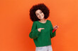 © khosrork - Smiling woman with Afro hairstyle wearing green casual style sweater holding and pointing at red envelope in her hand, romantic letter. Indoor studio shot isolated on orange background.