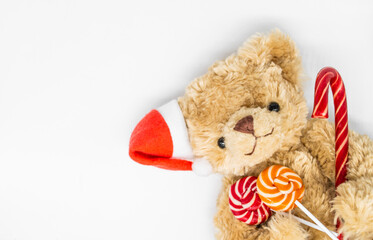 A stuffed toy Teddy bear in a red Santa Claus hat on one ear, holding candy cane and two striped lollipops in its paws. White background, copy space. Concept of Christmas