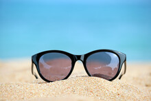 Closeup Of Black Protective Sunglasses On Sandy Beach At Tropical Seaside On Warm Sunny Day. Summer Vacation Concept.