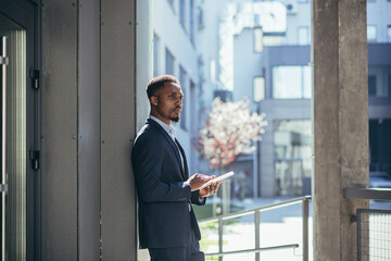 Wall Mural - Young african american businessman in formal business suit standing working with tablet in hands on background modern office building outside. Man using smartphone or mobile phone outdoors city street