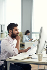 Wall Mural - Confident young bearded business analyst comparing paper and online data while working in office