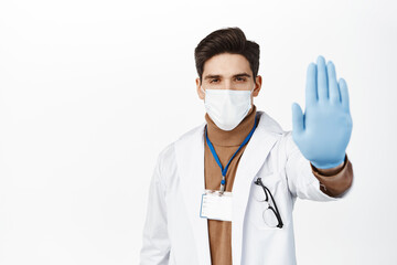 Wall Mural - Healthcare worker in face mask from covid 19, wearing rubber gloves, showing stop no gesture, taboo and prohibition sign, standing over white background