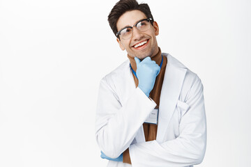 Wall Mural - Smiling doctor in glasses and medical gloves thinking, standing thoughtful, looking pleased up at logo, standing in medical uniform over white background