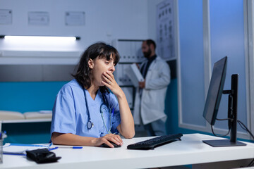 Exhausted nurse using computer on desk while falling asleep, working late. Medical assistant looking at monitor and feeling burnout after overtime work at night in healthcare office.