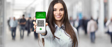 Green pass covid-19 vaccination certificate concept, smiling woman show digital vaccine certification qr code on smartphone screen, crowd of people in background, reopen and go back to new normal