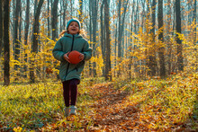 A Joyful Child Girl In A Jacket And Cap With A Ball In Her Hands Running Through An Autumn Forest. In The Background Is Green Grass, Yellow Leaves And Tree Trunks. The Concept Of A Happy Childhood.
