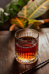 Faceted glass of whiskey with cocktail spoon on a wooden table against the background of exotic plants.