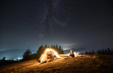 Wall Mural - Man sitting in brightly lit tent in the middle of mountain hill at night, playing at favorite guitar for his lover girl on the background of starry sky, forests and contour of mountain peaks.