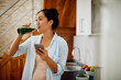 Happy Asian woman drinks healthy smoothie while using mobile phone in kitchen.