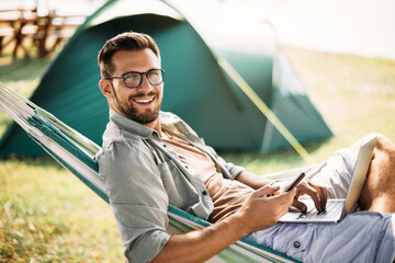 Wall Mural - happy man uses smartphone and laptop while camping in nature and looking at camera.