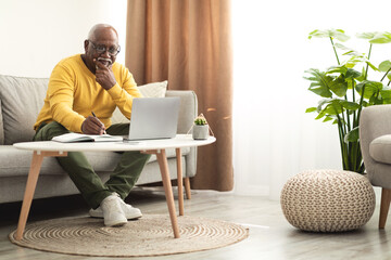 Wall Mural - Senior Black Male Using Laptop Working Online Taking Notes Indoor