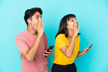 Young mixed race couple holding mobile phone isolated on blue background shouting with mouth wide open to the lateral