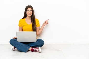 Young woman with a laptop sitting on the floor pointing finger to the side