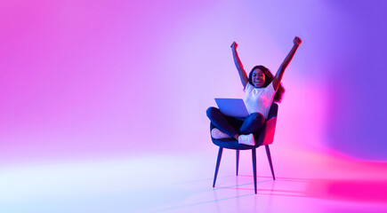 Wall Mural - Joyful young black woman sitting in armchair with laptop, celebrating huge online win or success in neon light, banner