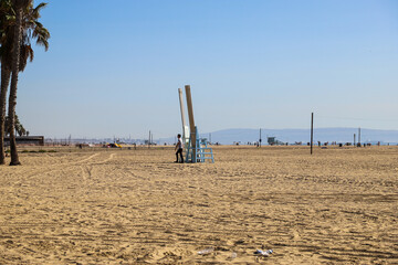 a couples standing next to two giant chairs at the beach with silky brown sand and vast blue ocean water with lush green palm trees and blue sky at Santa Monica Beach in California USA