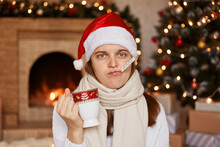 Sad Unhealthy Woman Wearing Santa Claus Hat, Being Wrapped In Warm Scarf, Sitting Near Christmas Tree, Measuring Temperature With Thermometer In Mouth, Holding Cup With Tea.