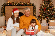 © sementsova321 - Indoor shot of happy family giving Christmas present to little cute dark haired girl in red wrapped gift box, posing in decorated living room near xmas tree and fireplace.