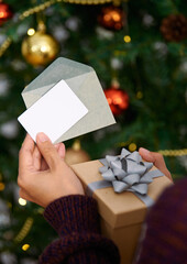 Hand holding mockup christmas greeting card and gift box for invitation design on christmas tree background.