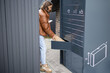 Young caucasian woman putting parcel to cell of automatic post terminal. Smiling girl holding smartphone and looking at camera on city street. Concept of smart delivery. Idea of shipping and logistics