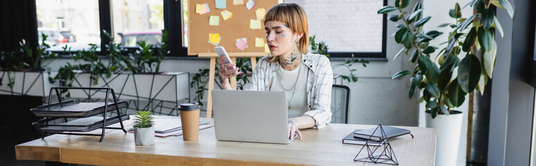 young and stylish businesswoman using cellphone while working near laptop in office, banner