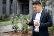 © rh2010 - Asian businessman with coffee cup calling on smartphone on city street. Concept of modern successful man. Idea of remote and freelance work. Person wearing suit