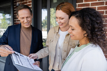 middle aged realtor holding folder with loan application near happy and interracial lesbian couple