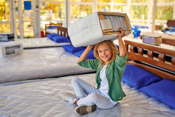 Wall Mural - Beautiful child cute girl sitting on the bed and holding a cutaway mattress sample in the store. Purchase of bedding.