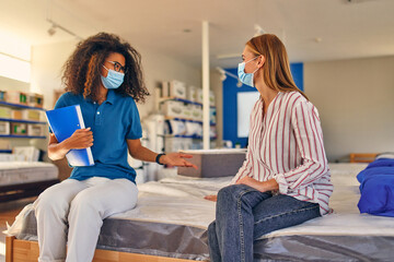 Wall Mural - African American Saleswoman consultant in protective medical mask advising a woman in a store of mattresses, beds and pillows. Quarantine during the covid pandemic.