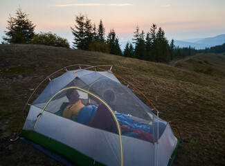 Wall Mural - Couple of travelers, who just woken up in their white tent, watching on the morning colors of summer sky. Magic sunrise over mountain peaks. Overnight campers in tent on mountain hill.