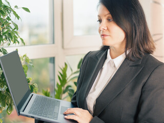 businesswoman holding laptop. adult woman wearing suit holding silver laptop in office on window background