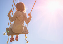 Back View Of Carefree Happy Young Girl On Swing In Sunlight Against Blue Sky. Copy Space For Text Or Design.
