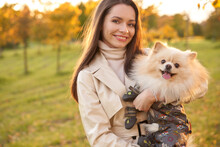 Young Beautiful Woman With Long Straight Hair Walking In Park With Pomeranian Spitz Dog. Elegant Woman In Bright Jacket And Blue Jeans In Autumn Park