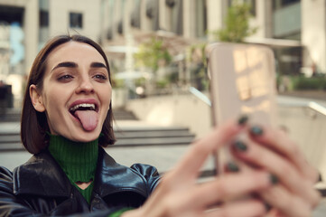 Close-up of cheerful funny joyful woman holding mobile phone in outstretched hands and grimacing, showing tongue, taking self-portrait while walking around city