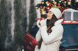 © polinaloves - Happy beautiful young woman wearing knitted sweater and woolen hat drinking hot chocolate near red car with Christmas tree on the top under snowfall.