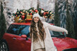 © polinaloves - Happy beautiful woman wearing knitted sweater and woolen hat standing near red car with Christmas tree on the top under snowfall.