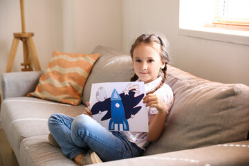 Wall Mural - Little girl with drawing of rocket sitting on sofa at home