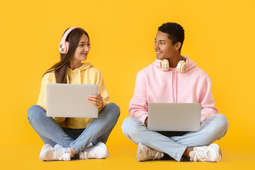 Wall Mural - Stylish young couple in hoodies using laptops on yellow background