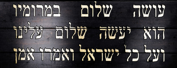 Wall Mural - Last lines of Jewish prayer Kaddish on black wooden background. Translation is: He who creates peace in His celestial heights, may He create peace for us and for all Israel, and say, Amen.