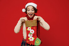 Young Surprised Fun Woman 20s Wear Santa Claus Christmas Red Hat Hold Look At Christmas Stocking Gift Sock Isolated On Plain Red Background Studio Portrait. Happy New Year 2022 Celebration Concept