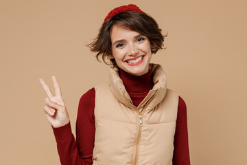 Poster - Young smiling happy fun cheerful friendly caucasian woman 20s wears red turtleneck vest beret showing victory sign isolated on plain pastel beige background studio portrait. People lifestyle concept.