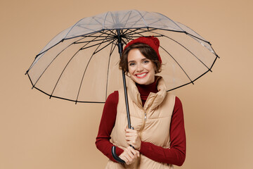 Poster - Young smiling happy woman 20s wears red turtleneck vest beret hold transparent umbrella look camera isolated on plain pastel beige background studio portrait. People lifestyle rainy weather concept.