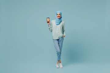 Wall Mural - Full body young arabian asian muslim woman in abaya hijab hold takeaway delivery craft paper brown cup coffee to go isolated on plain blue background People uae middle eastern islam religious concept