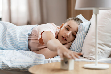 Wall Mural - people, bedtime and rest concept - sleepy girl with alarm clock awaking in bed at home