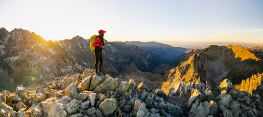 Wall Mural - Lady hiker with backpack standing on top of the mountain and enjoying valley view. Hiker meets the sunset. Adventure concept. 