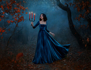 Fantasy woman queen runs in dark forest, hands hold vintage burning candlestick three candles. Blue velvet long medieval dress flies in wind. Gothic girl princess. Autumn nature night, bare trees fog