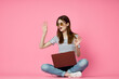 © VICHIZH - cheerful woman with laptop sitting on the floor on a pink background